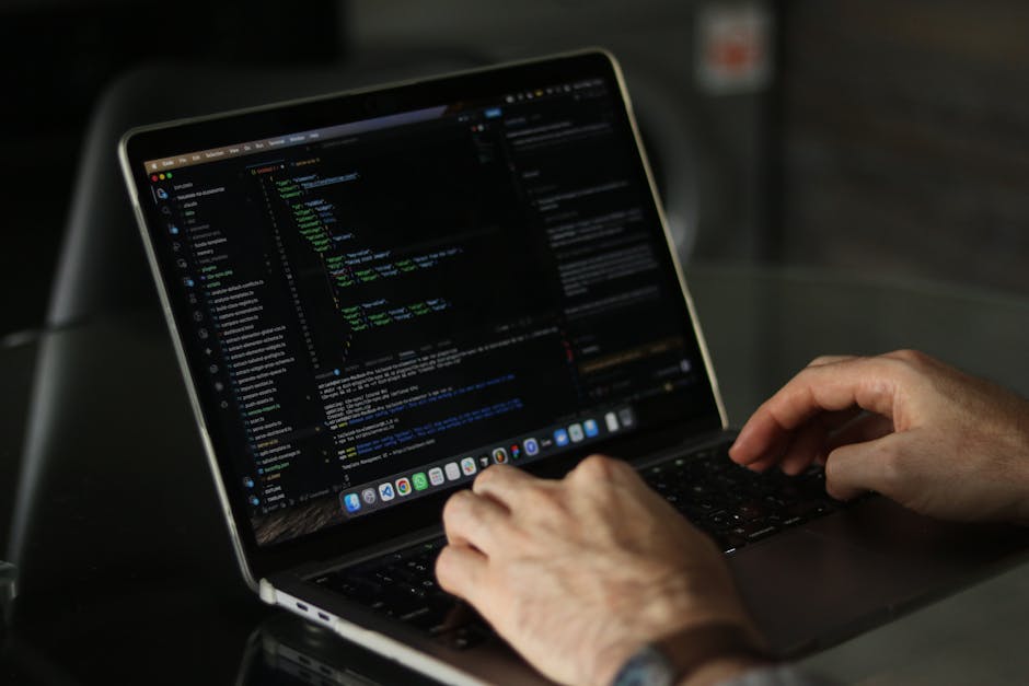 A close-up view of a person's hand typing on a laptop keyboard placed on a wooden desk. The laptop screen displays lines of code with colorful syntax highlighting, indicating a programming or web deve