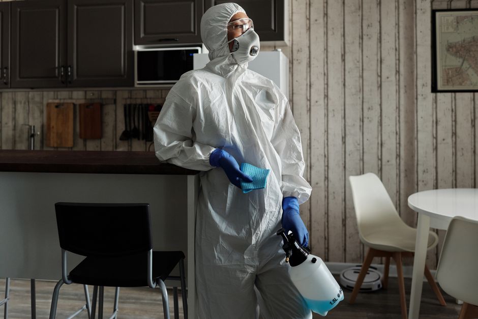 A professional cleaner dressed in full protective suit, gloves, and shoe covers performing deep cleaning on a light-colored carpet in a well-lit living room. The room features white walls, a window wi