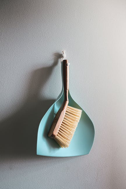 A light blue dustpan and long-handled brush hanging on a plain white wall, with the brush's bristles facing downward and the handle resting inside the dustpan. The scene depicts a clean, well-organize
