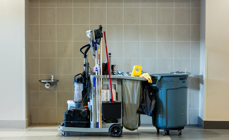 A street cleaner dressed in bright orange reflective safety gear, including a jacket, trousers, and cap, is standing on a paved sidewalk at night. The cleaner holds a broom with a wooden handle and a